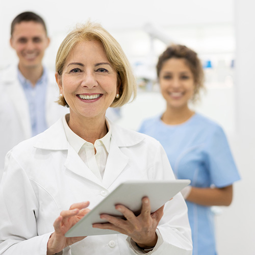 a dentist holding a tablet smiling at the camera, their team smiling in the background behind them